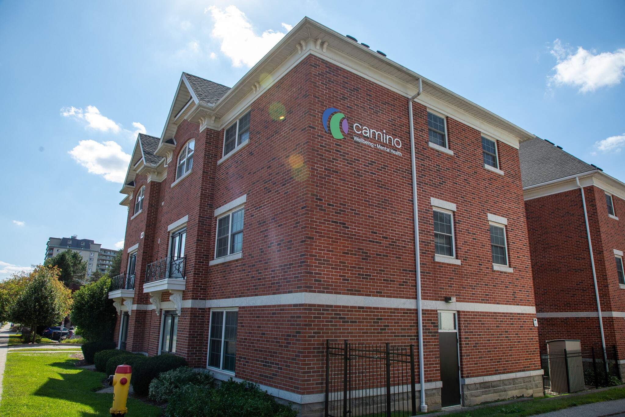 Three-story red brick building with Camino sign on the side, surrounded by grass and shrubs on a sunny day.