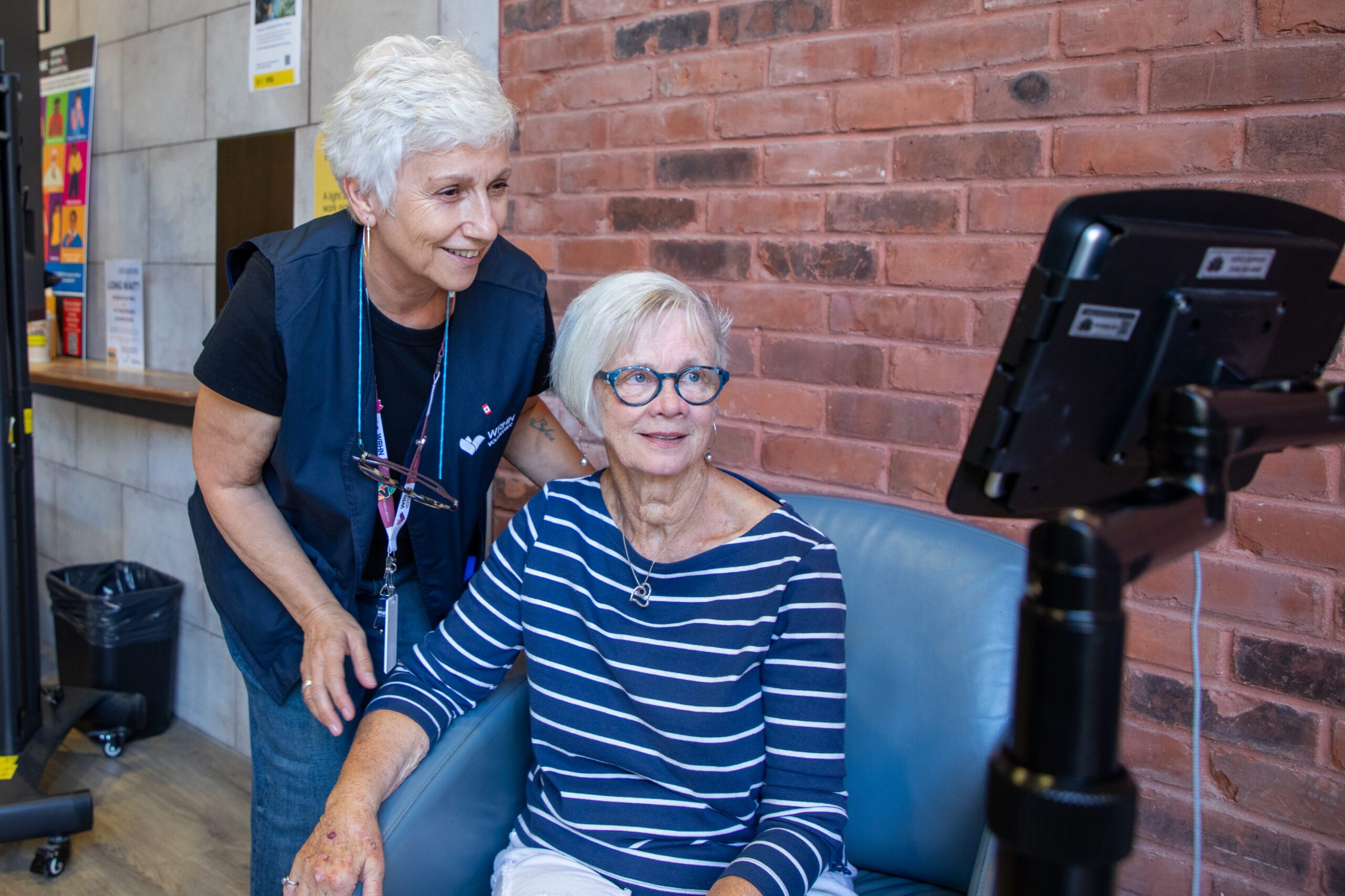 Two older women sit and stand together, smiling at a tablet mounted on a stand in a brightly lit indoor space.