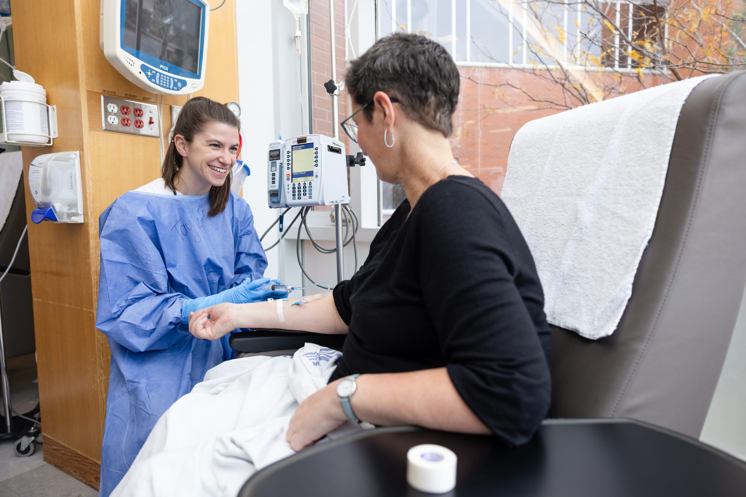 A healthcare worker in PPE administers an IV to a seated patient in a medical treatment room.