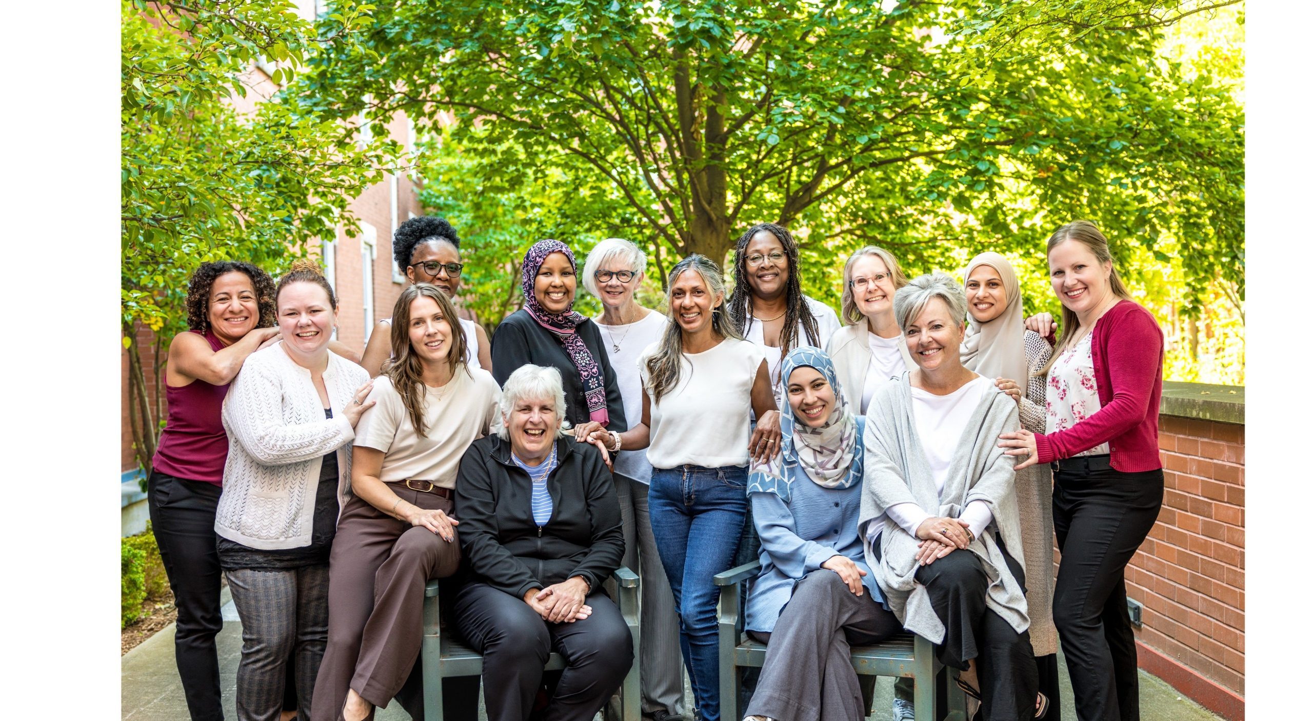 A group of fifteen women pose and smile together outdoors in front of trees and a brick wall.