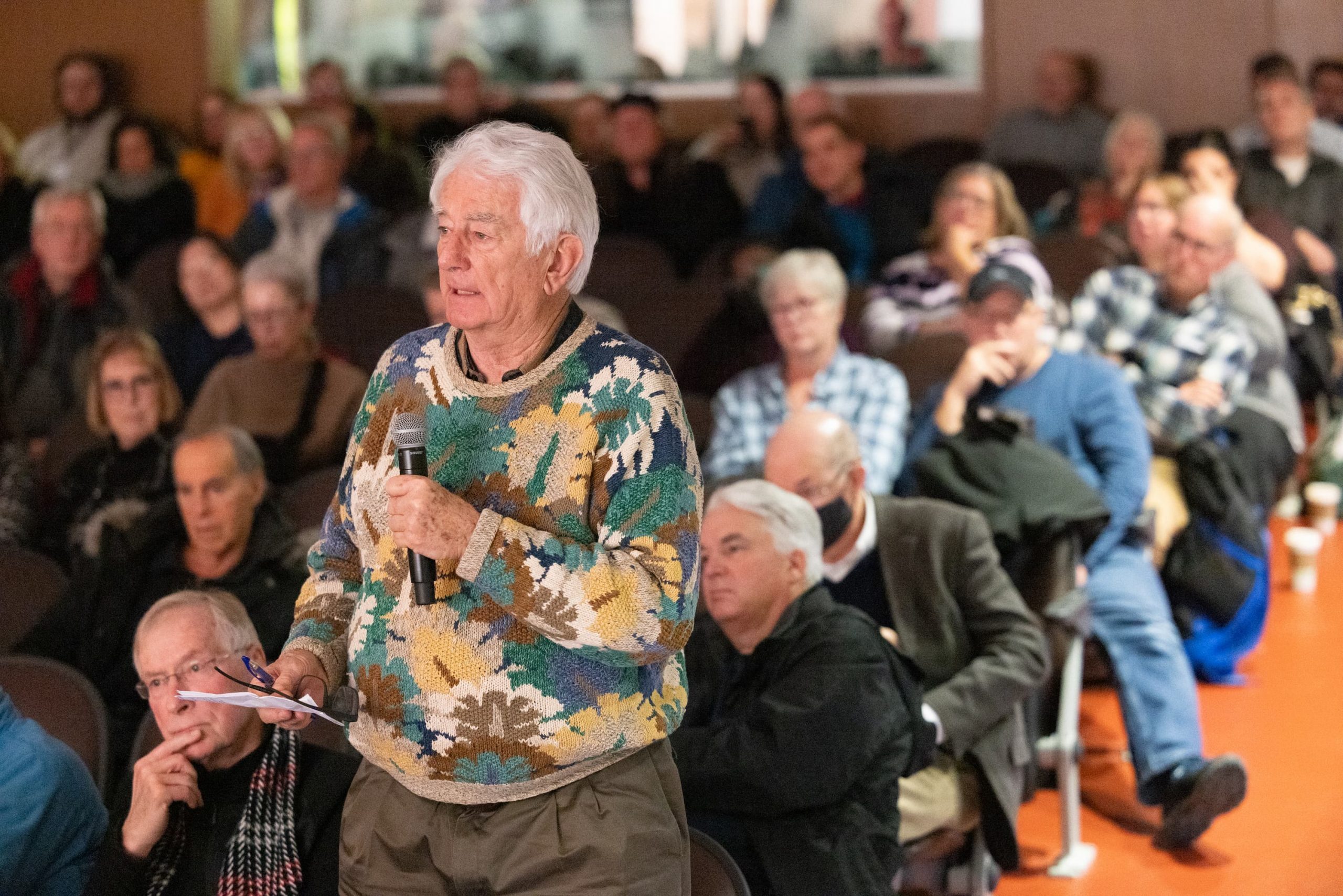 An elderly man in a patterned sweater holds a microphone and speaks to a seated audience in a crowded room.