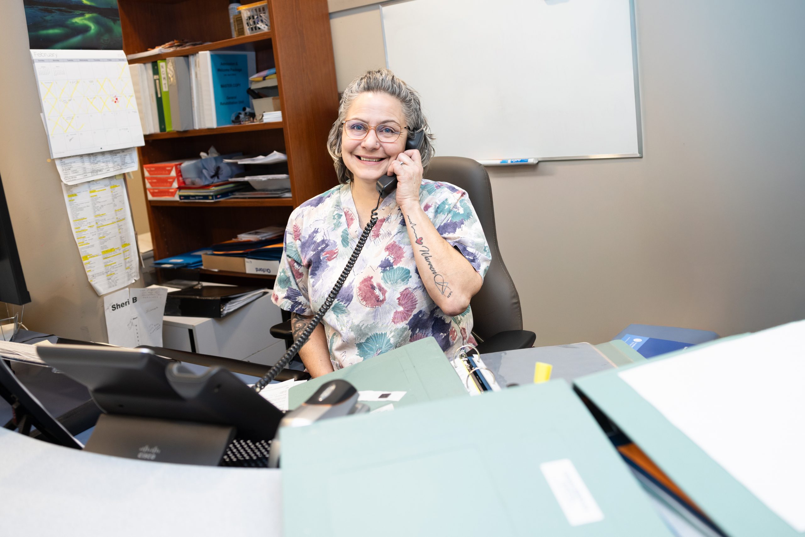A woman wearing scrubs sits at a desk, smiling and talking on the phone in an office setting.