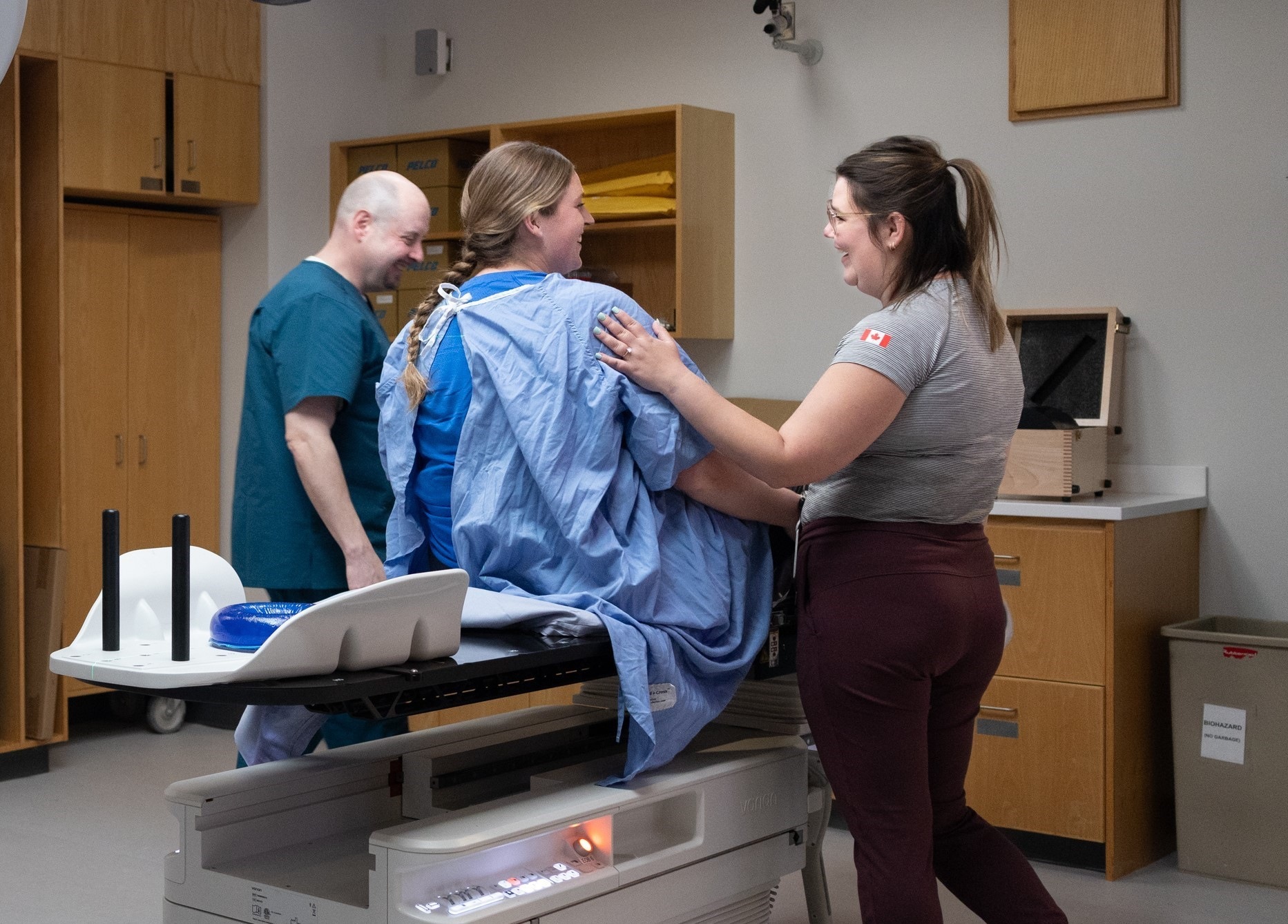 A woman in a hospital gown sits on an exam table with two medical professionals standing beside her.