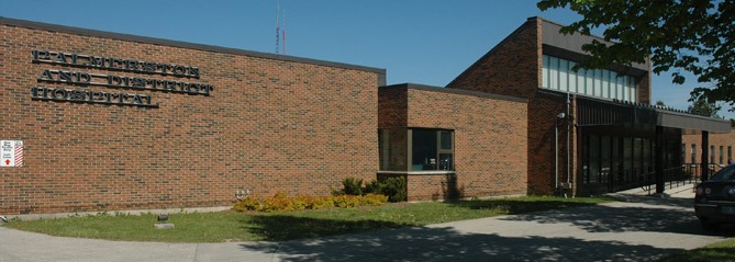 Brick building with "Palmerston and District Hospital" sign above entrance, surrounded by pavement and greenery.