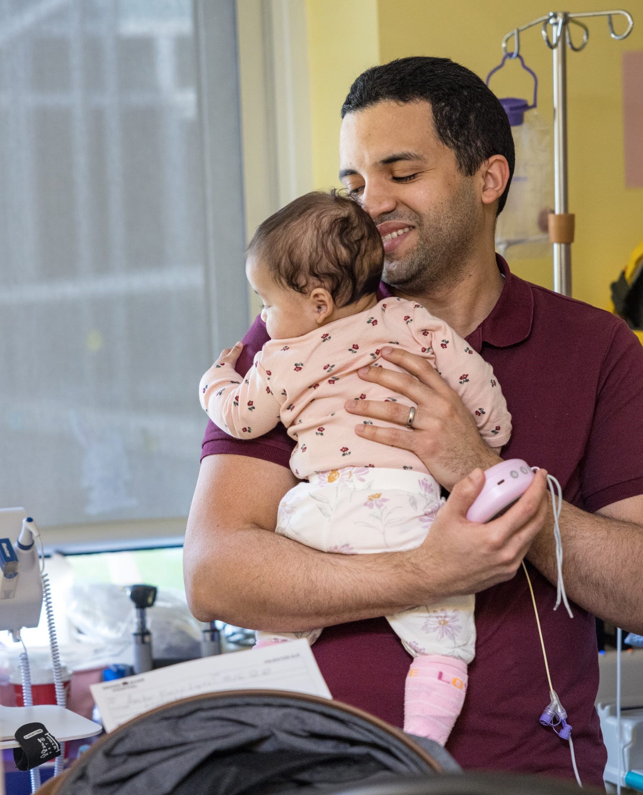 A man holds a baby in a hospital room, both facing away from medical equipment and monitors.