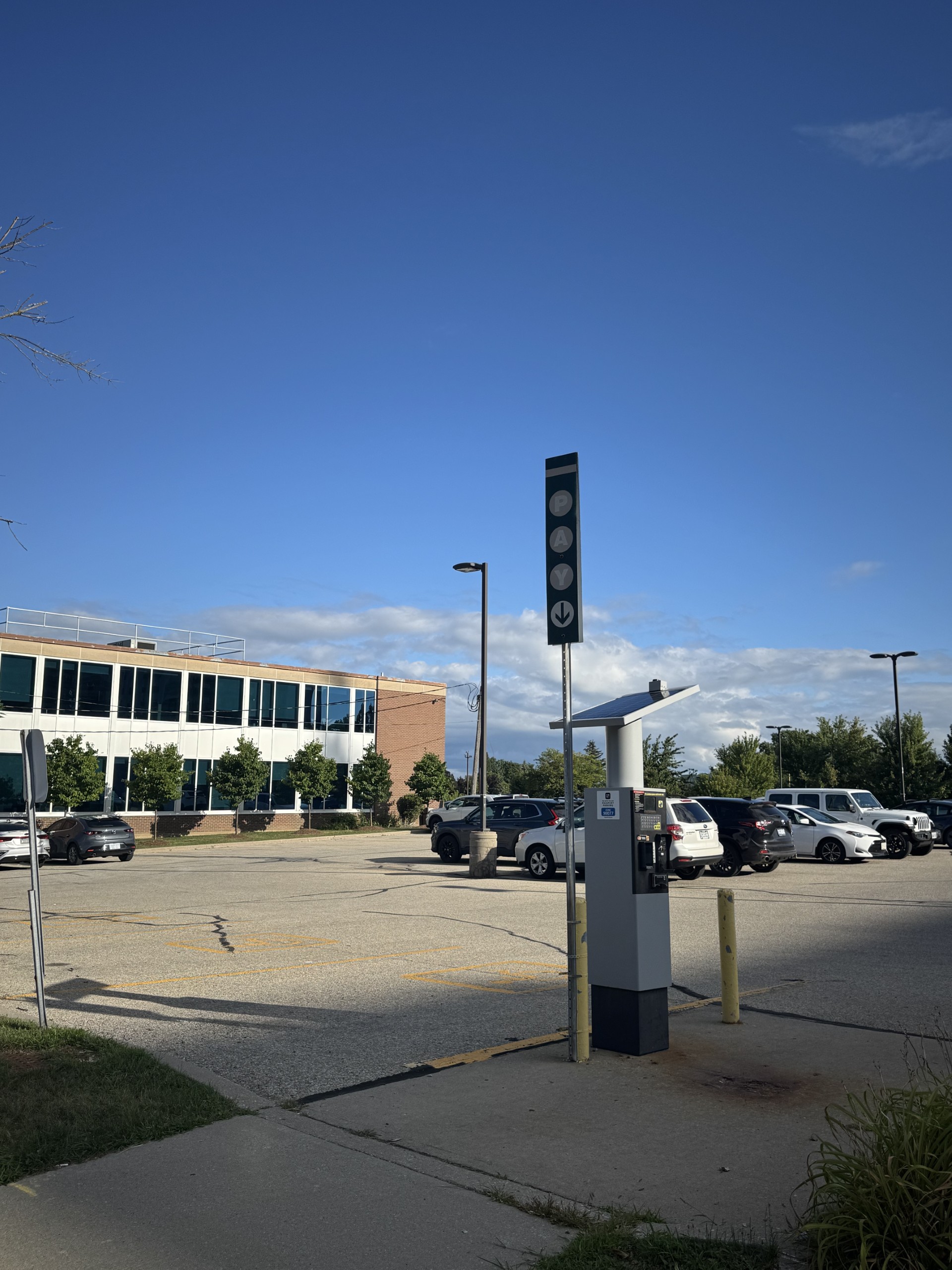 A parking lot with several parked cars, a pay station, and a nearby building under a clear blue sky.