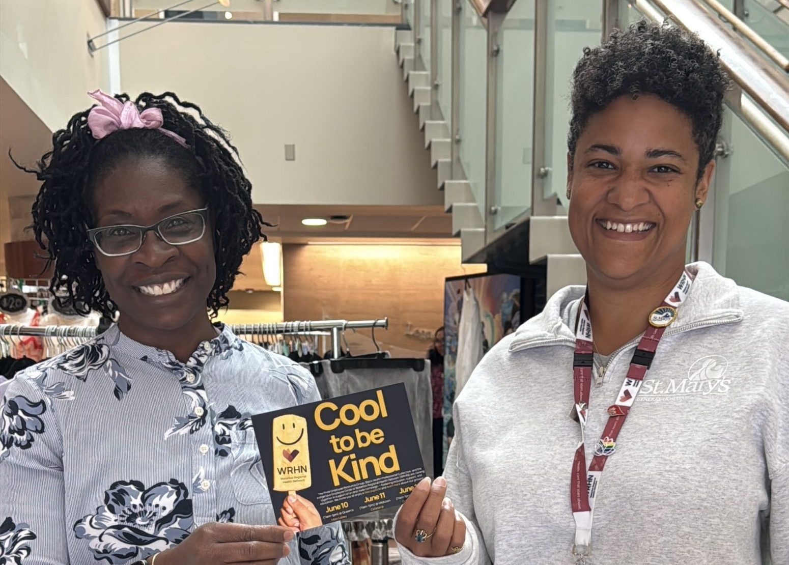 Two women stand behind a table with "Cool to be Kind" signs and stickers at an indoor event.