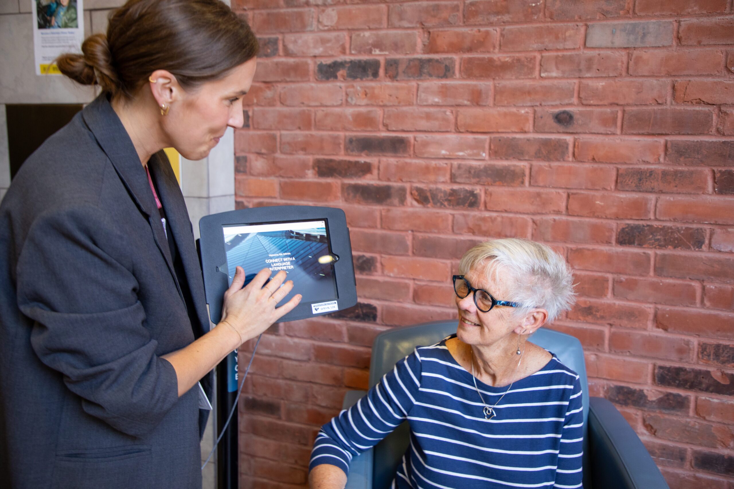 A woman shows information on a tablet to an older woman seated in a chair against a brick wall.