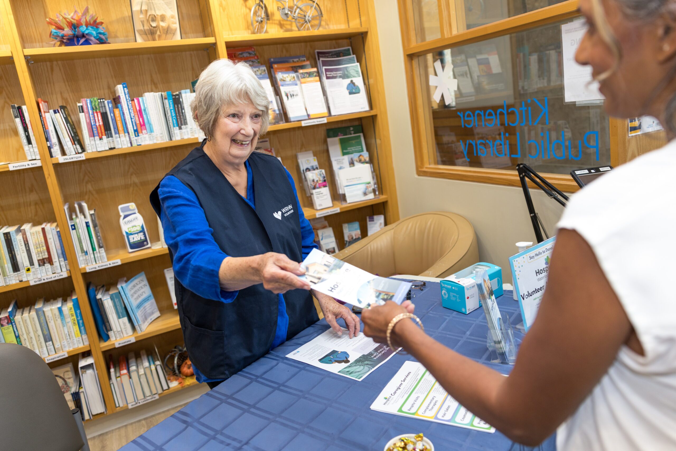 Photo of a woman with grey hair handing a brochure to a woman in a white t-shirt.