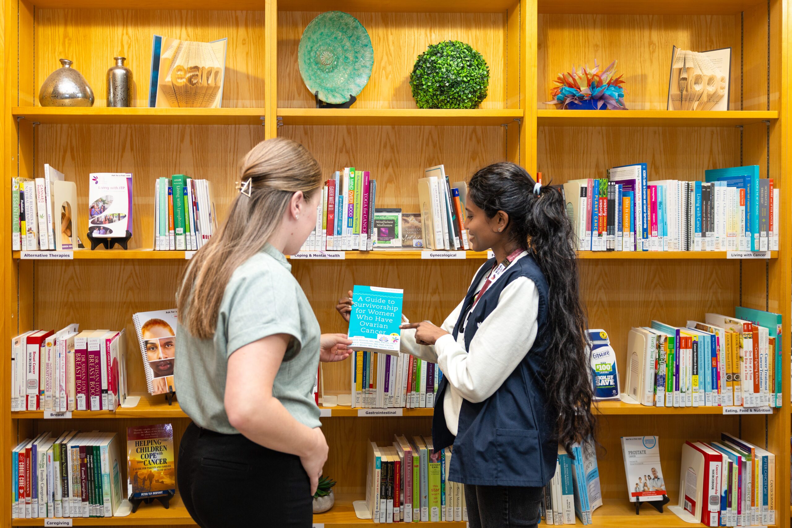 Two women stand in a library, looking at a book together in front of shelves filled with books and decorative items.