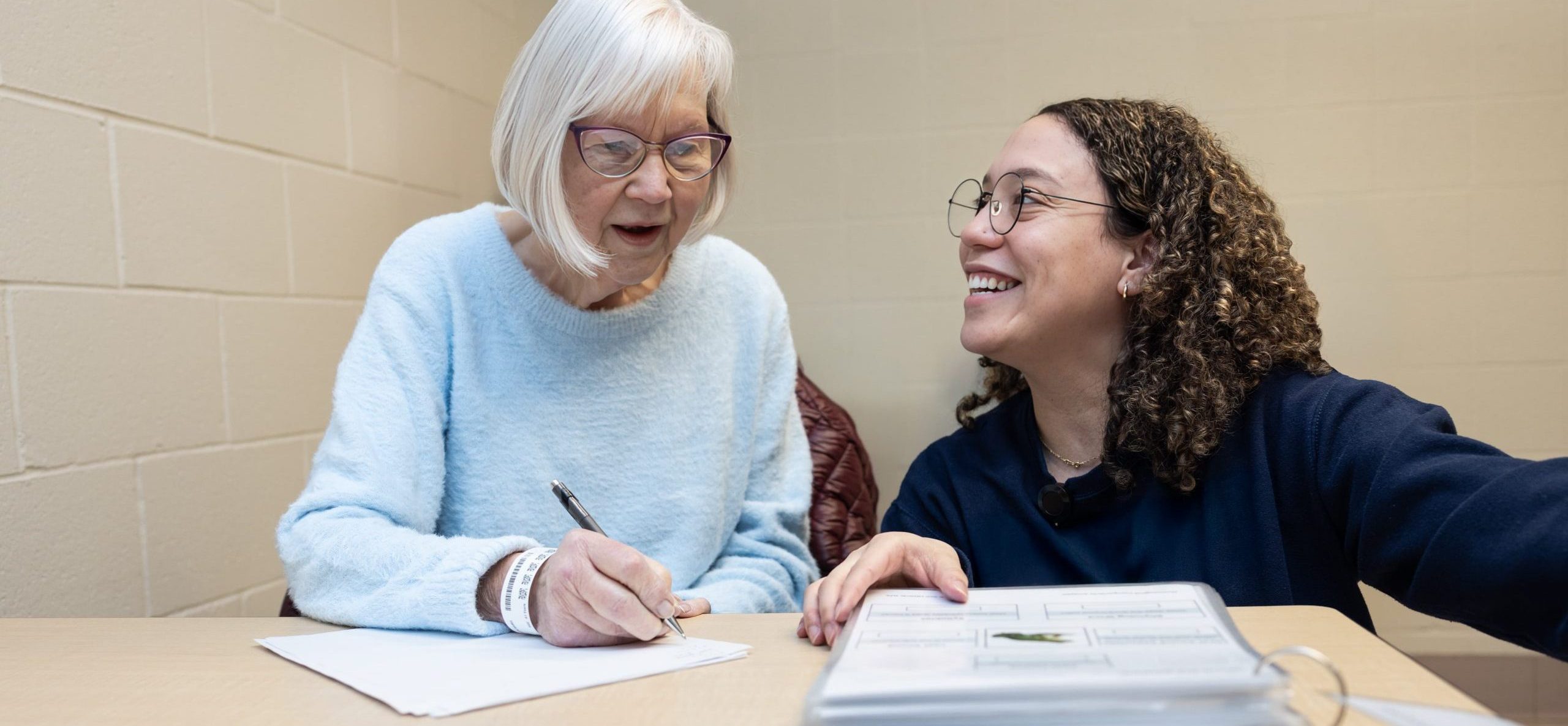 An older woman writes on paper while a younger woman with a binder sits beside her and smiles.