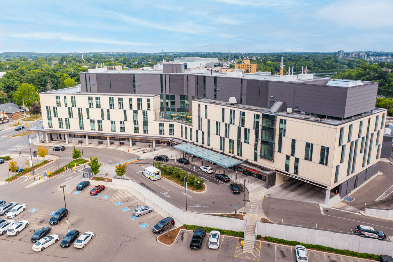 A large modern hospital building with parked cars in the lot, surrounded by greenery under a blue sky.