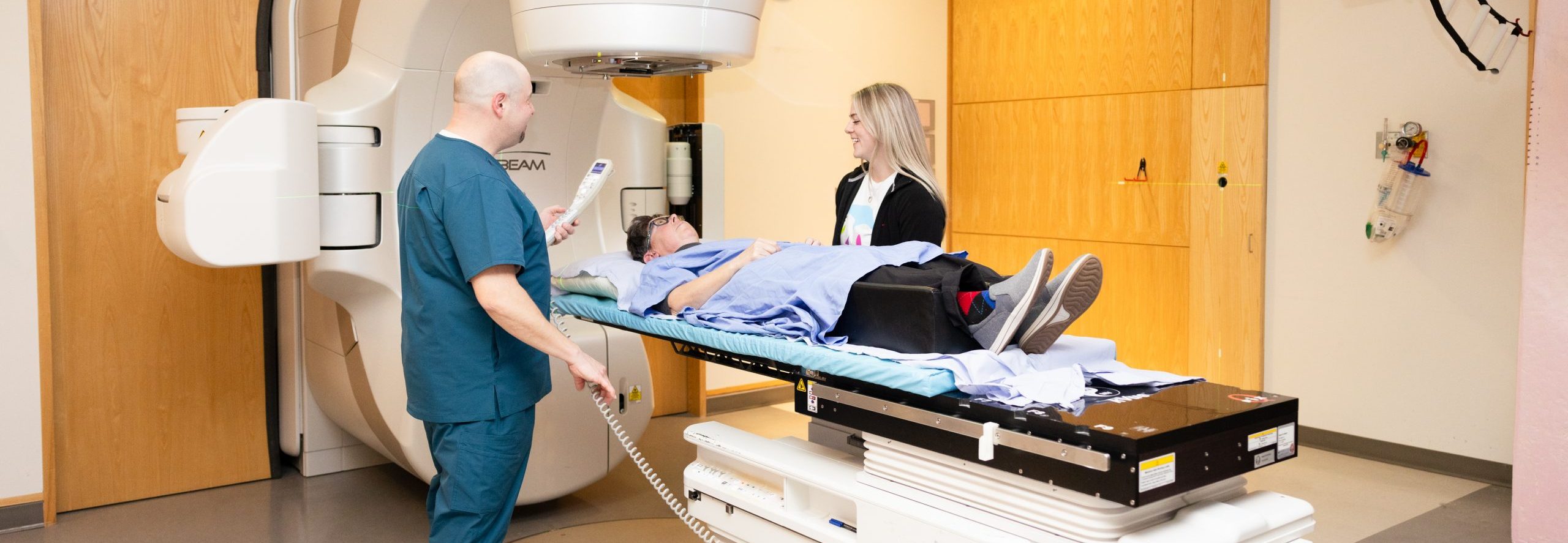 A patient lies on a treatment table as two medical professionals prepare equipment in a radiation therapy room.