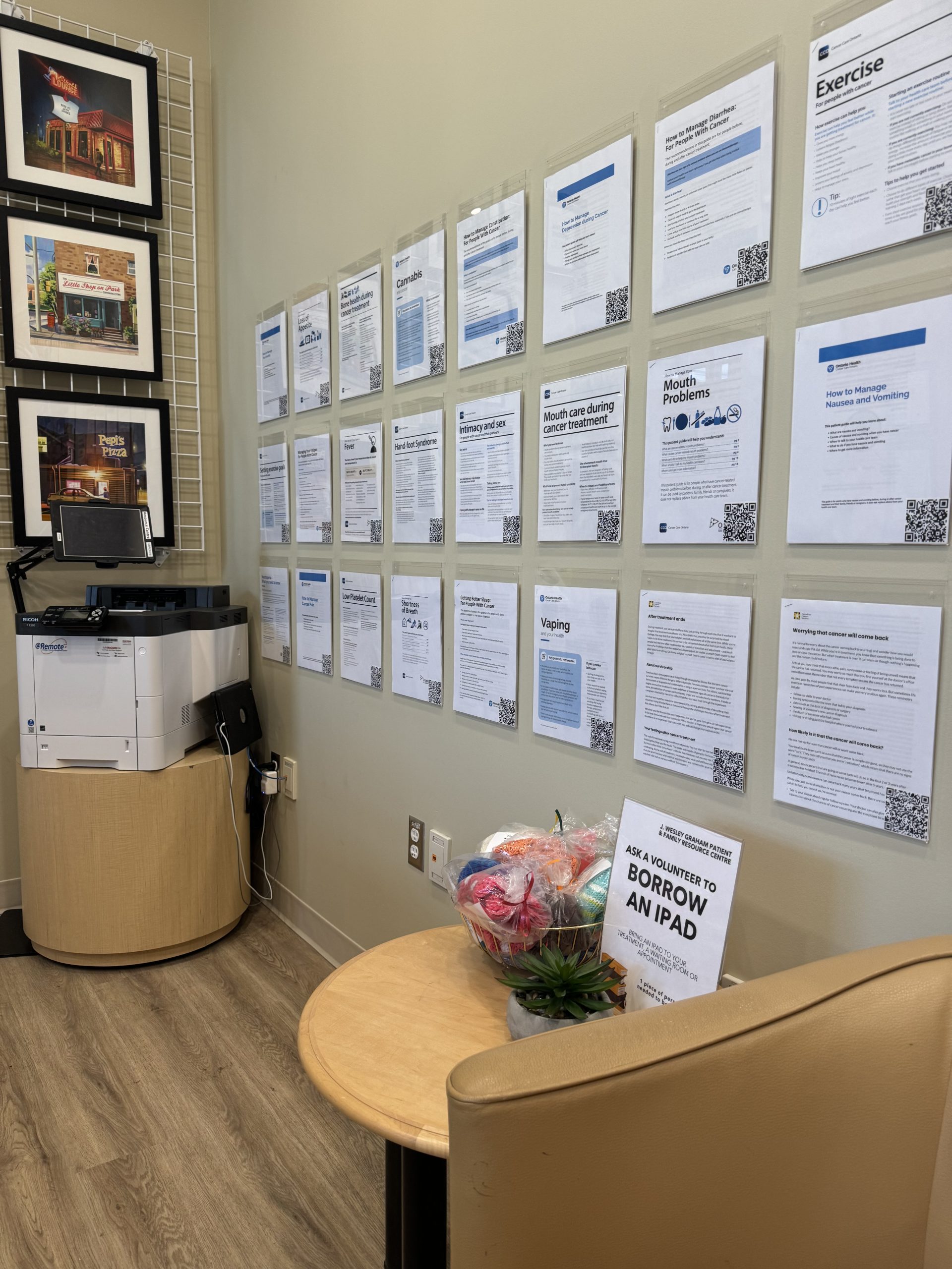 A wall with printed information sheets, a table with a gift basket, and a printer in a waiting area.