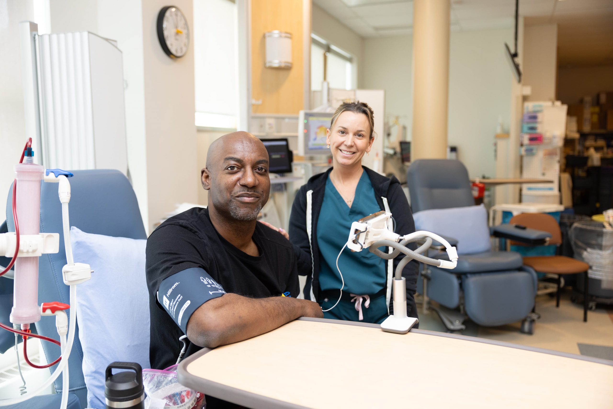 A patient sits next to medical equipment in a clinic while a healthcare worker stands nearby, both smiling at the camera.