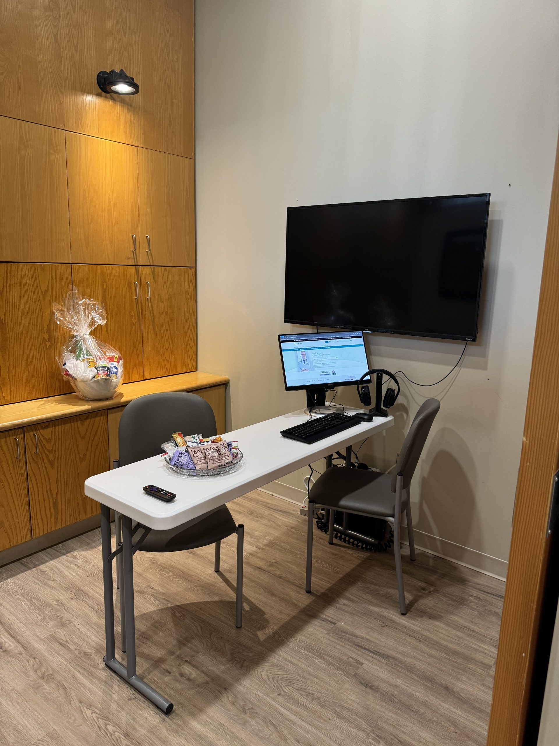 Small office with a desk, two chairs, a computer, a wall-mounted monitor, and a basket on a cabinet.