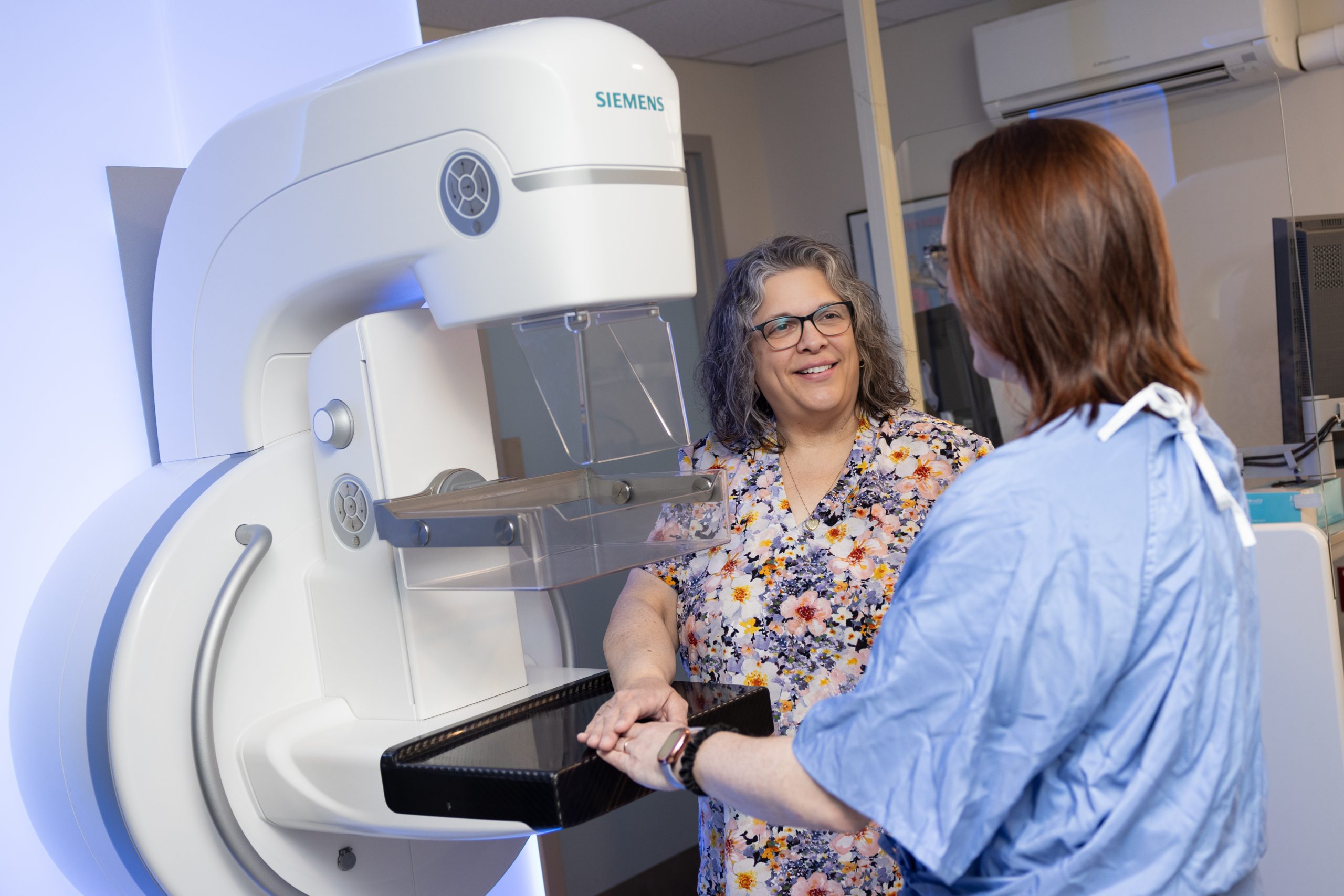 Two women stand by a Siemens mammography machine; one explains the procedure while the other listens attentively.