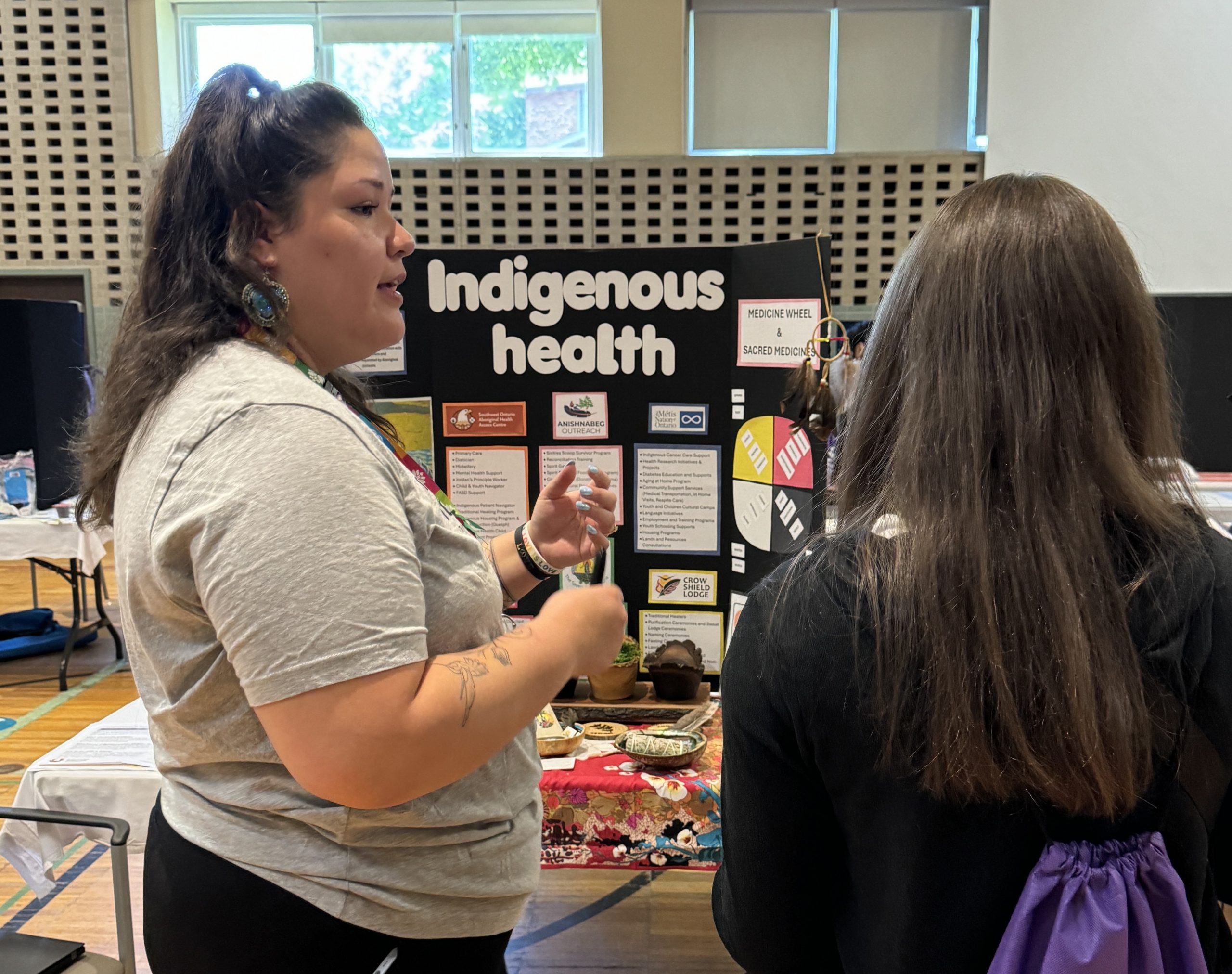 A woman speaks to a visitor at an informational booth about Indigenous health inside a community center.