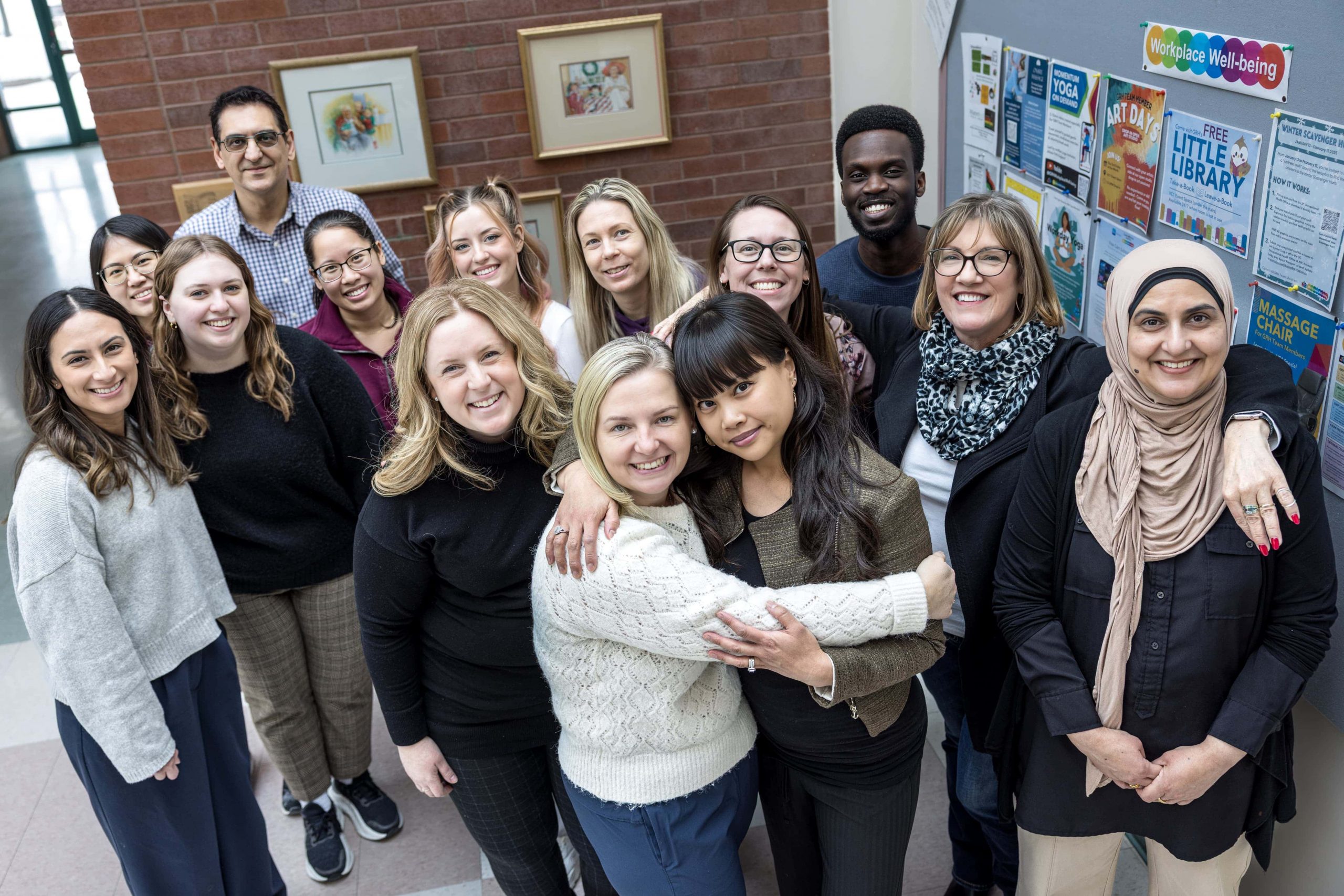 A diverse group of adults stands indoors, smiling at the camera in front of a bulletin board with community notices.