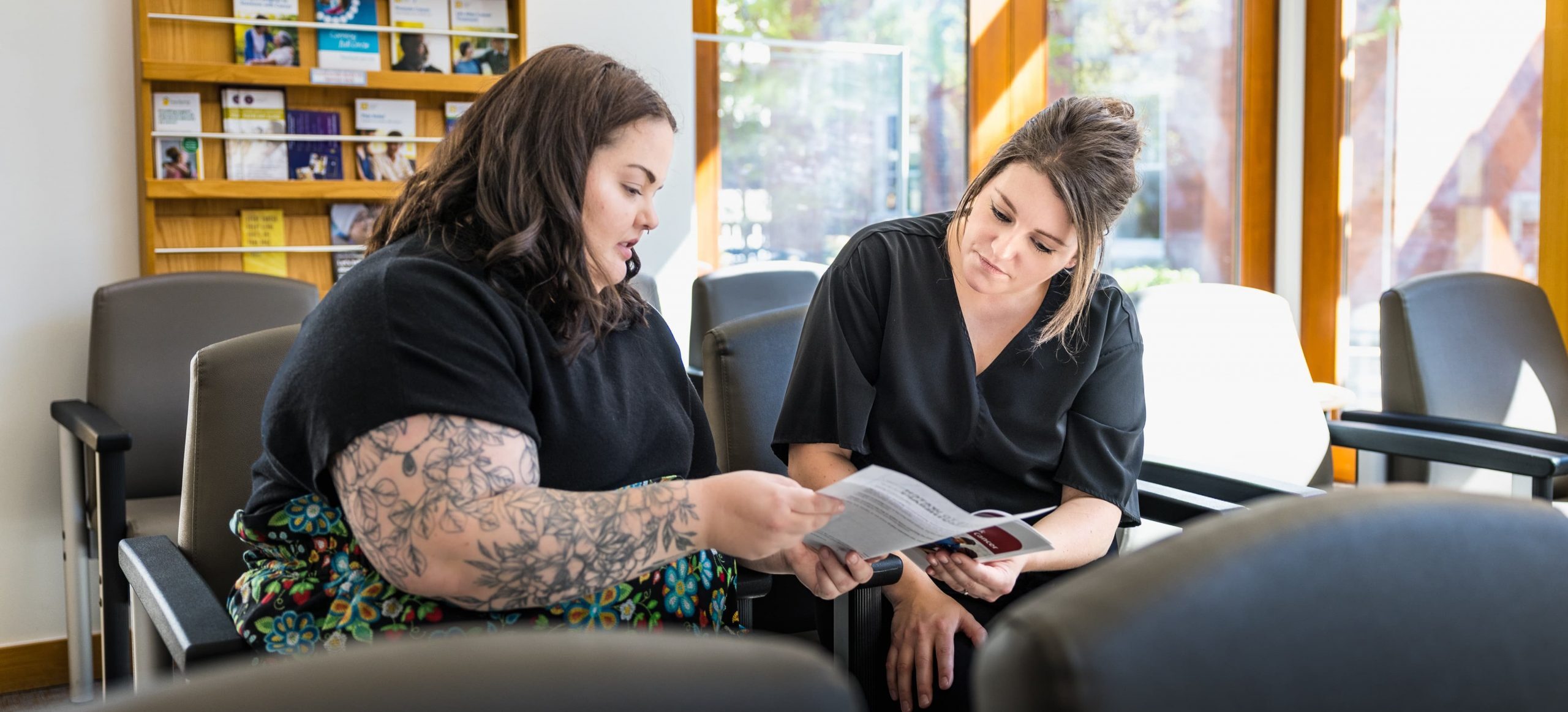 Two people sit in a waiting room, reviewing a document together. Shelves with brochures are visible in the background.