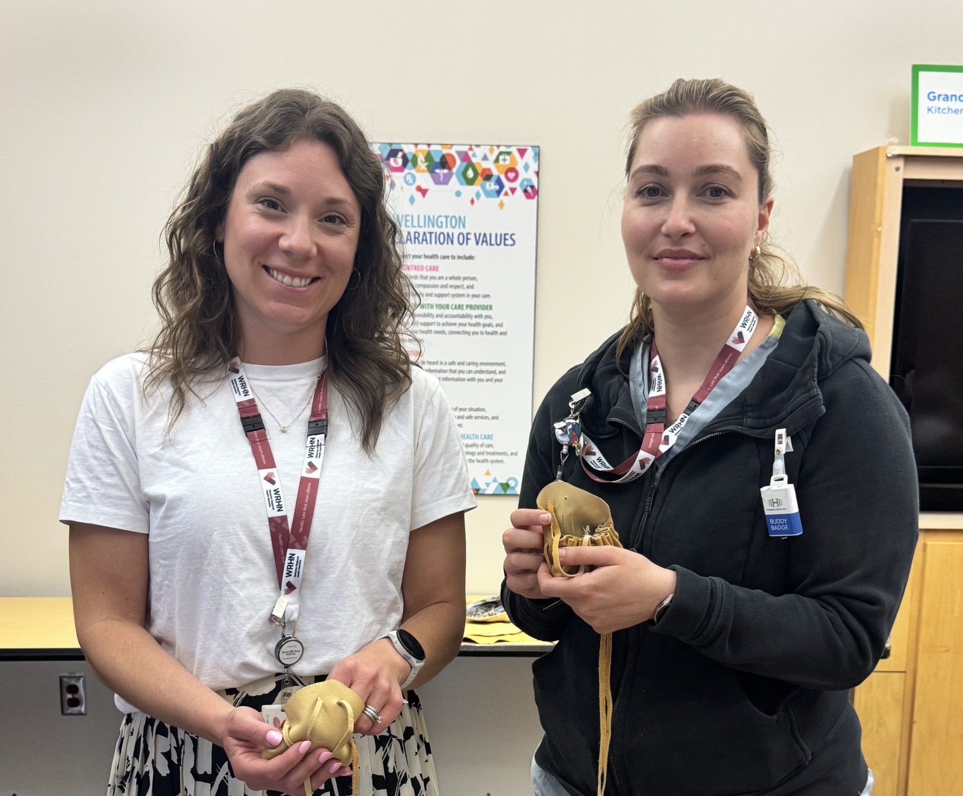 Two women stand indoors holding small wood carvings, with brochures and bowls of natural items displayed on a table in front of them.