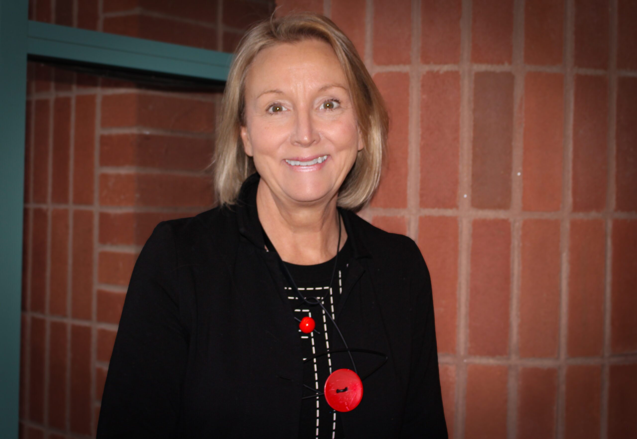 A woman with blonde hair and a black jacket stands indoors, smiling at the camera with a blurred background behind her.