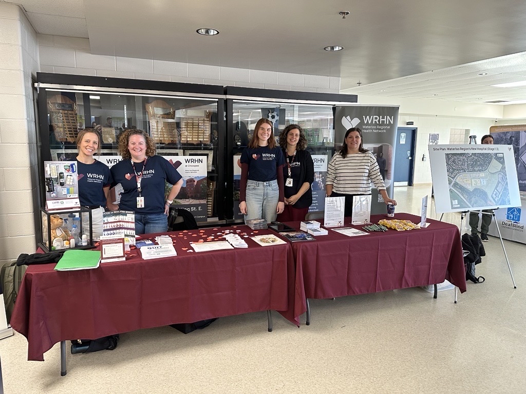 Five people stand behind a maroon-covered information table with brochures, snacks, and WRHN banners in a hallway.