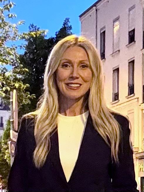 Une femme aux longs cheveux blonds, vêtue d'un blazer noir et d'un haut blanc, se tient debout à l'extérieur près de bâtiments au crépuscule.
