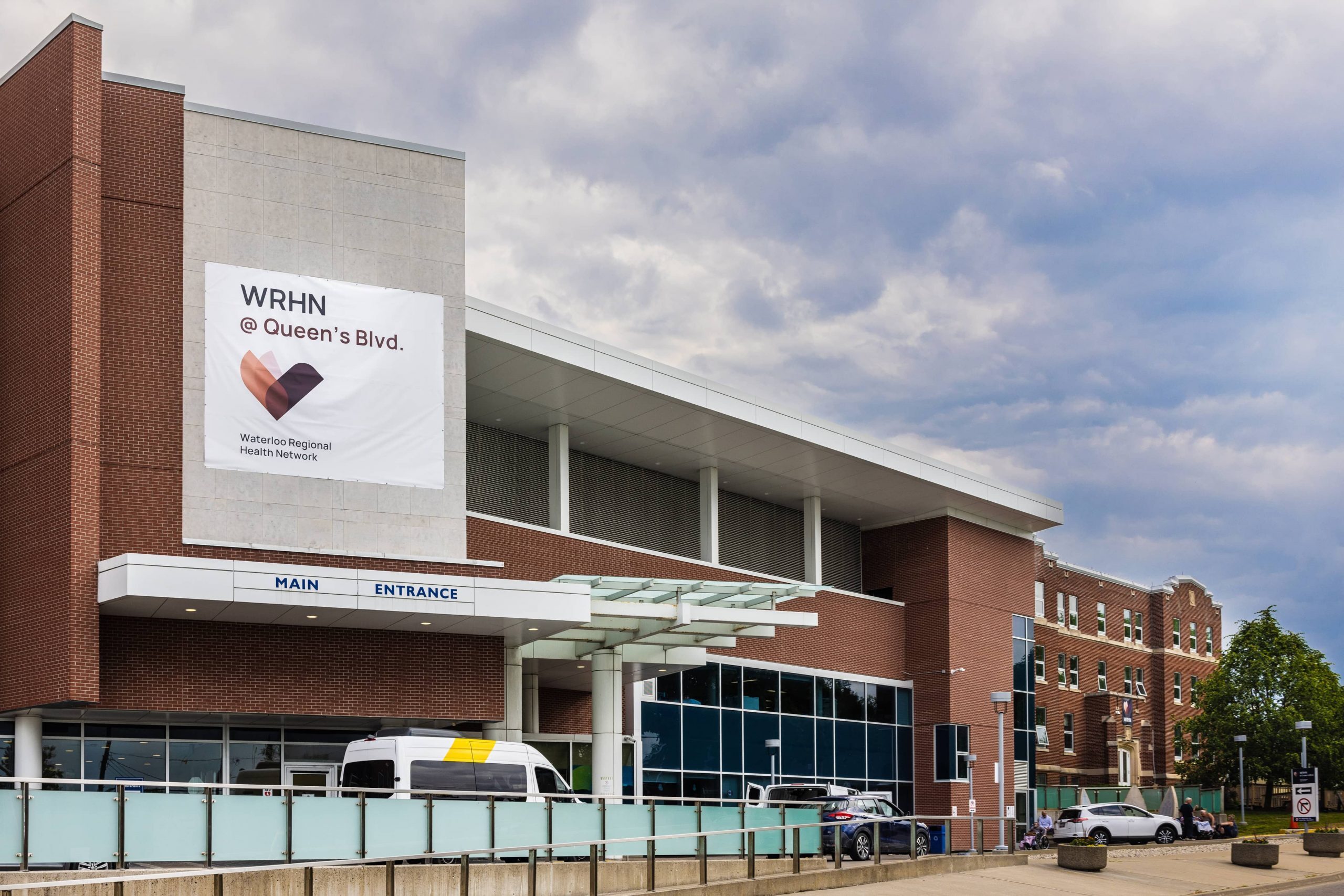 A modern hospital building with a sign reading “WRHN @ Queen’s Blvd.” above the main entrance, cars parked outside.