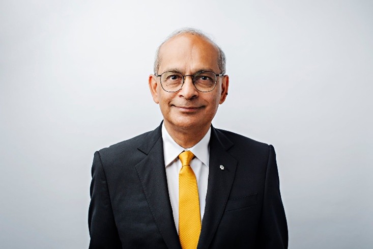 Portrait professionnel d'un homme en buste, portant des lunettes, un costume sombre, une chemise blanche et une cravate jaune vif. Il fait face à l'appareil photo avec un léger sourire, sur un fond gris clair. L'image présente un aspect soigné et formel, idéal pour un profil professionnel ou une biographie d'entreprise.