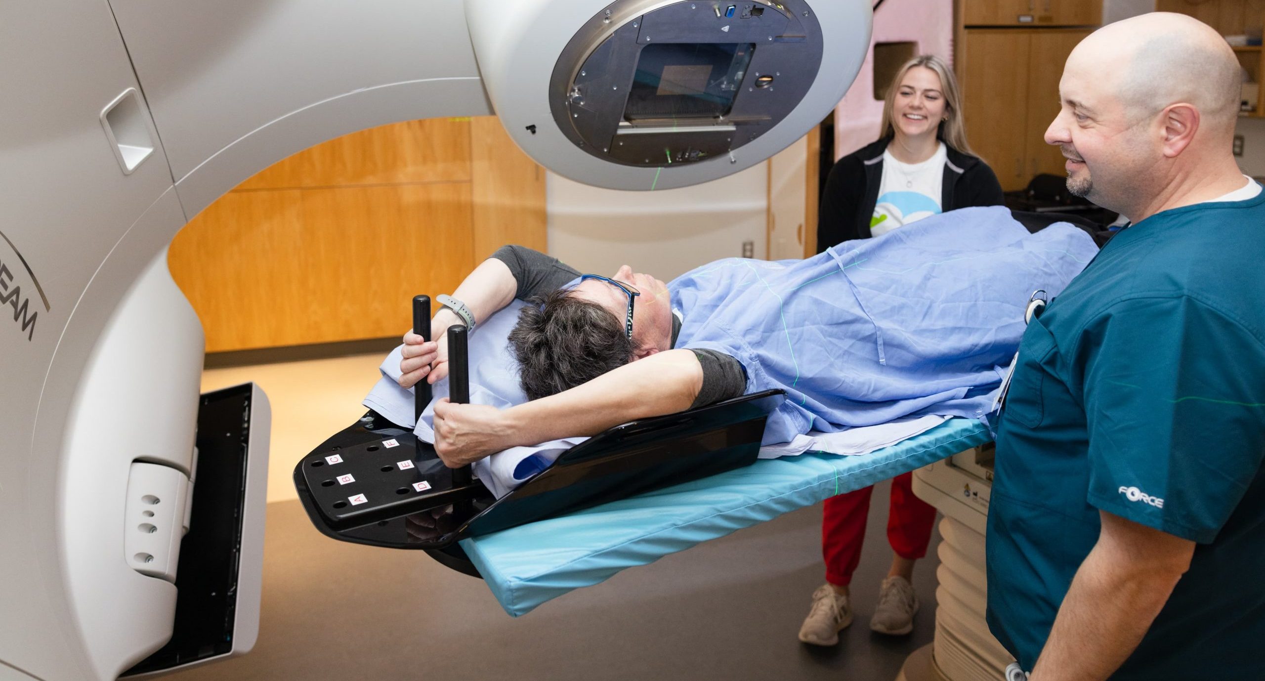 Ein Patient erhält eine Strahlentherapie, während er auf einem Behandlungstisch liegt und von zwei medizinischen Mitarbeitern in einer Klinik überwacht wird.