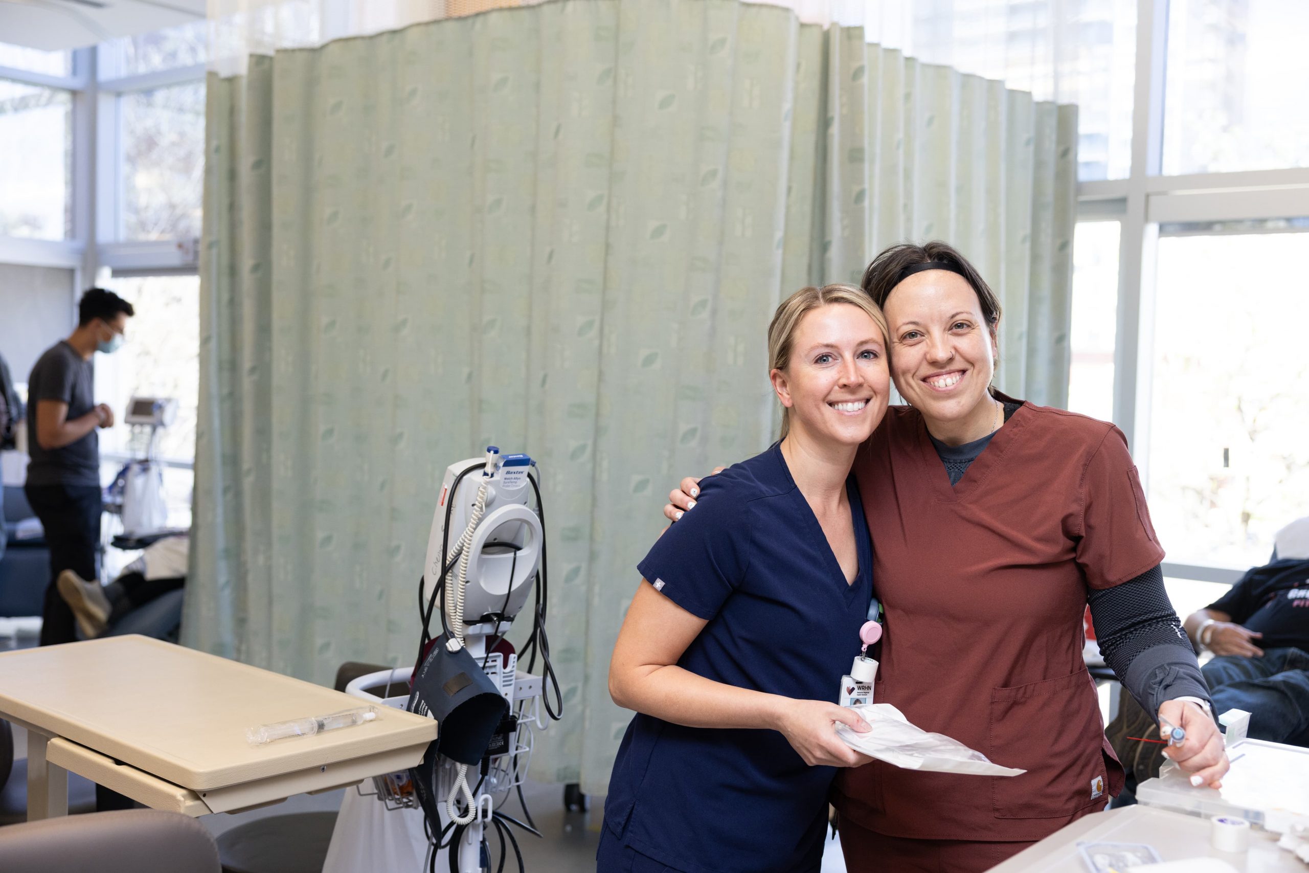 Deux infirmières en blouse sourient et posent ensemble dans une clinique médicale, avec du matériel et un paravent en arrière-plan.