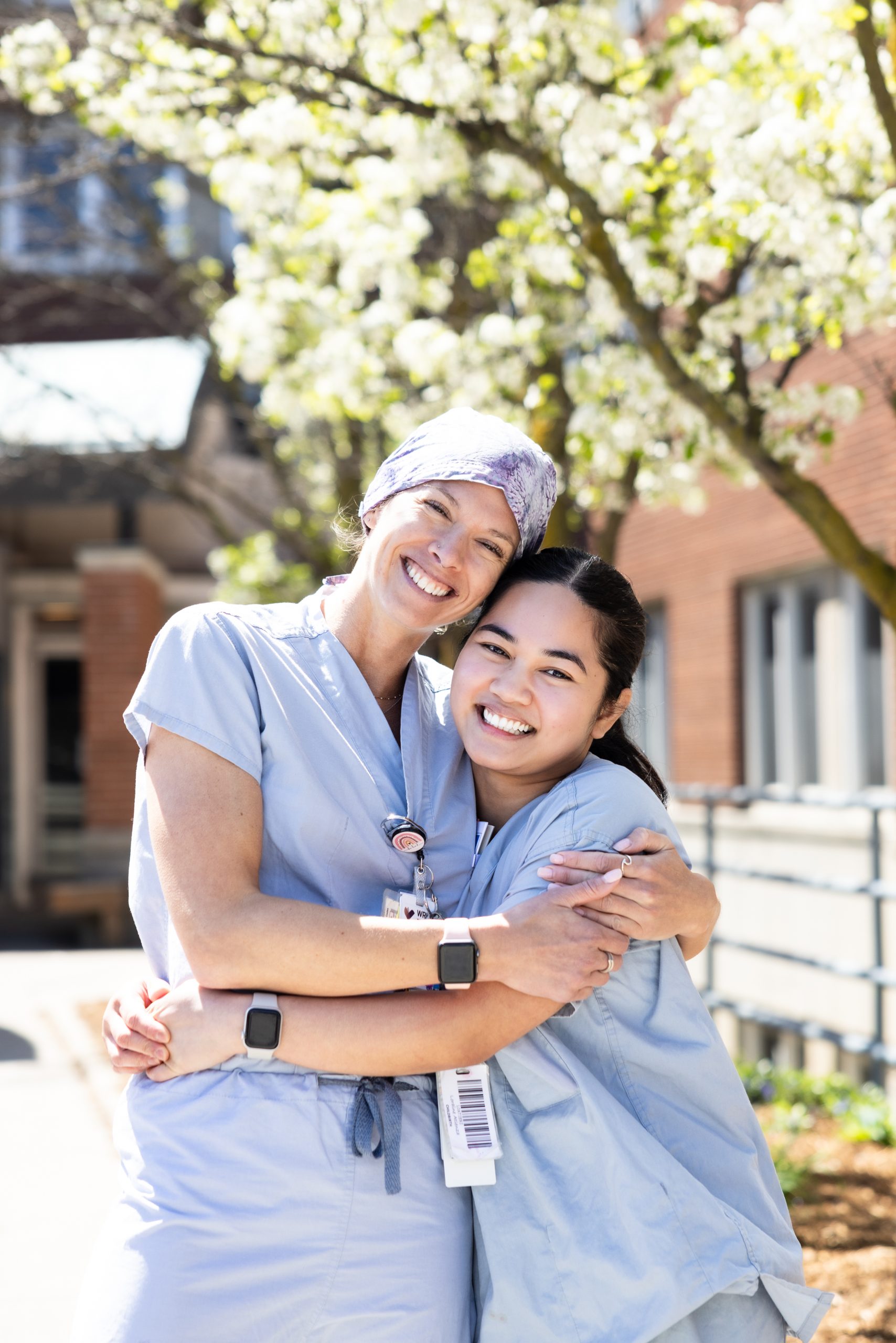 Two healthcare workers in scrubs smiling and hugging outside near blooming trees and a building.