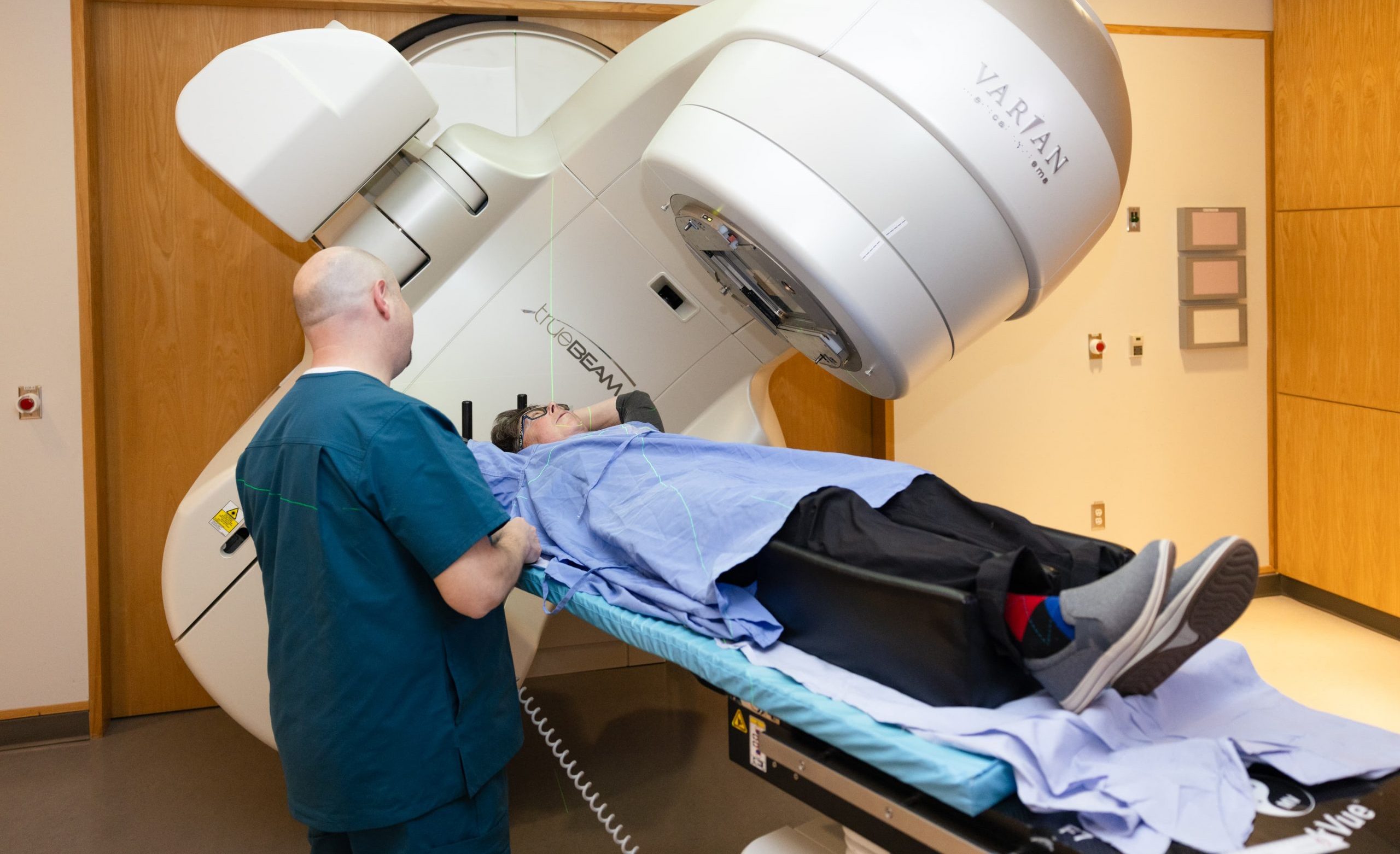 Ein Patient liegt auf einem Tisch unter einem großen medizinischen Gerät von Varian, während ein Mitarbeiter des Gesundheitswesens neben ihm steht.