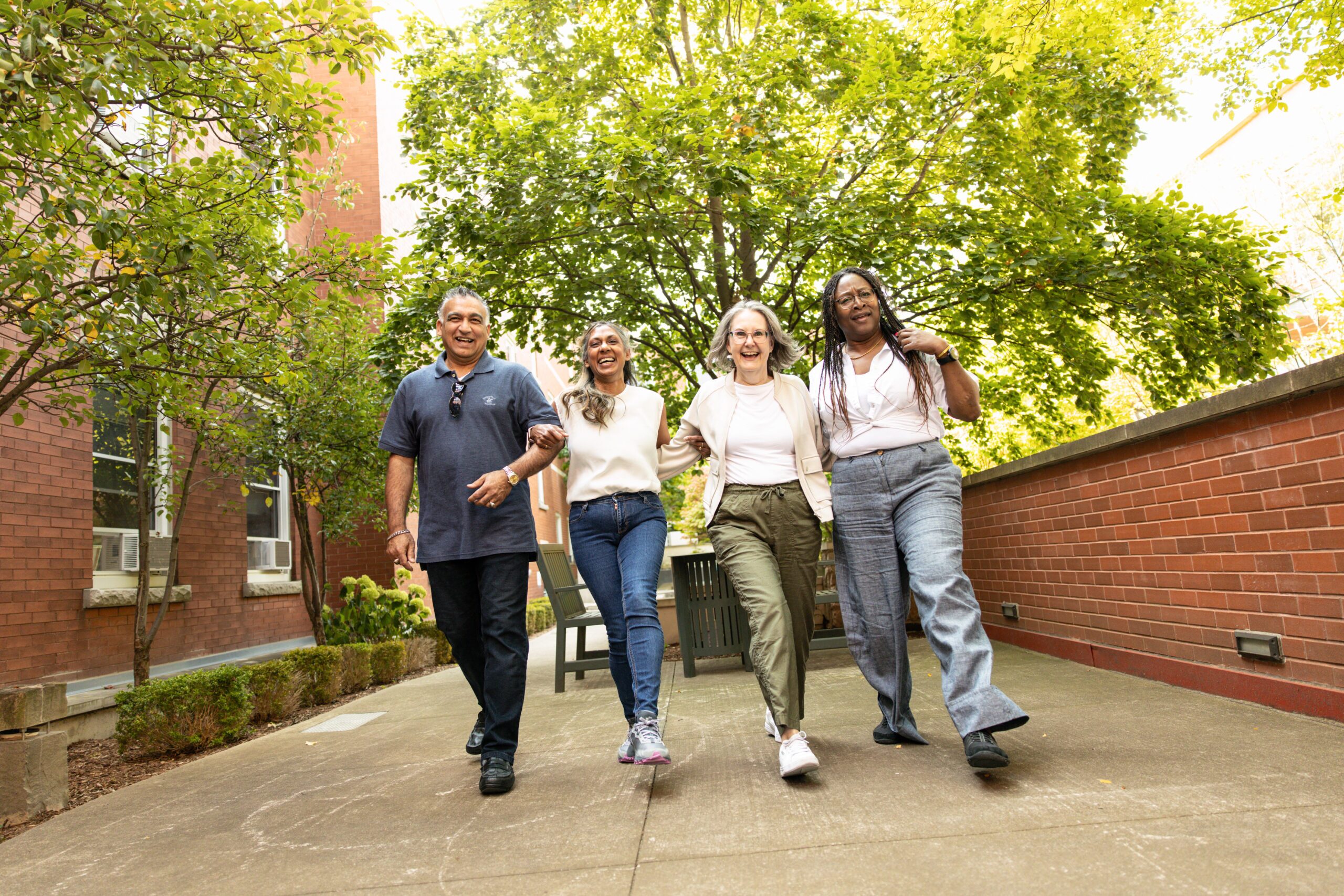 Four adults walk arm-in-arm down a tree-lined sidewalk in an outdoor urban setting on a sunny day.