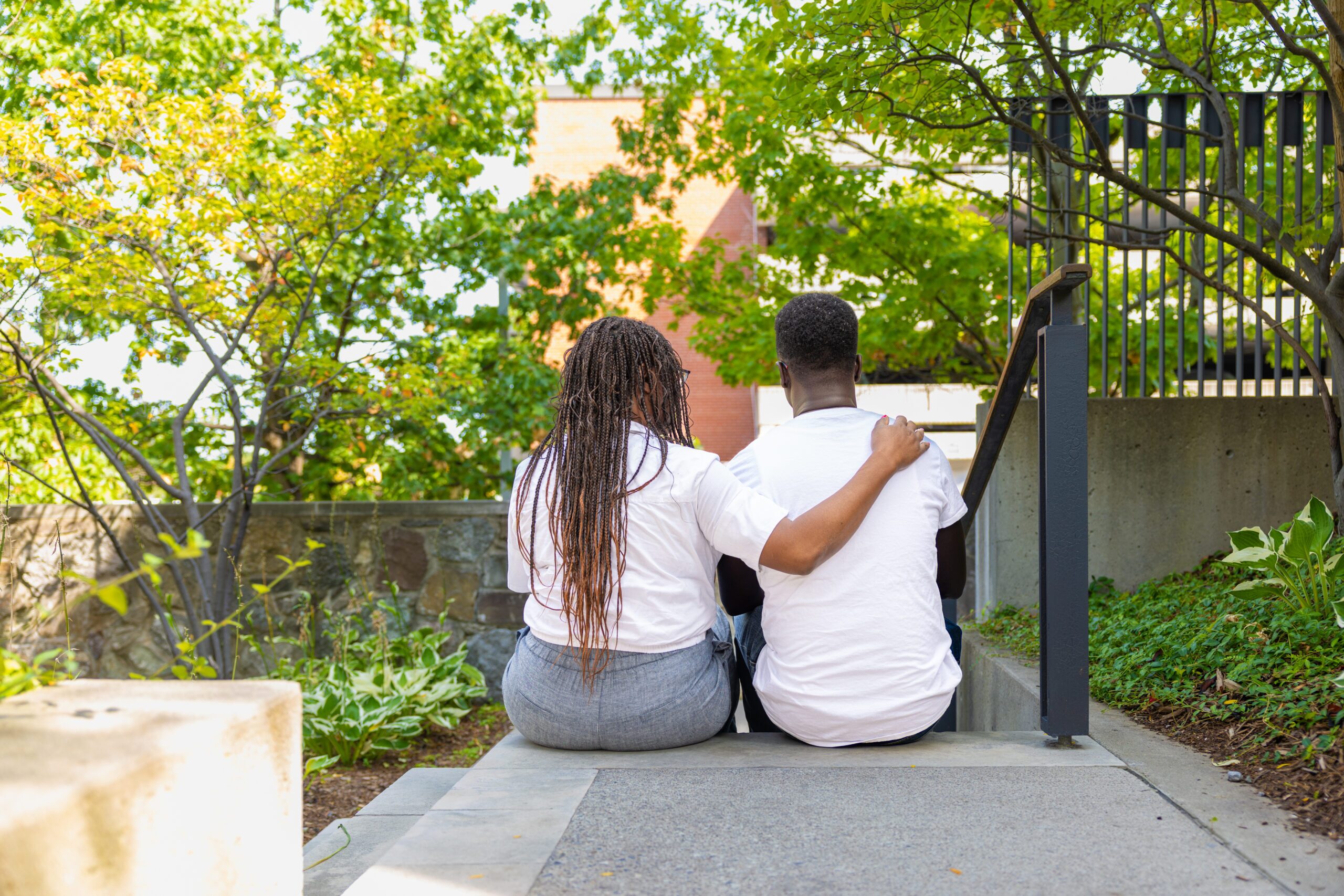 Two people sit side by side on outdoor steps, one with an arm around the other, both facing away from the camera.
