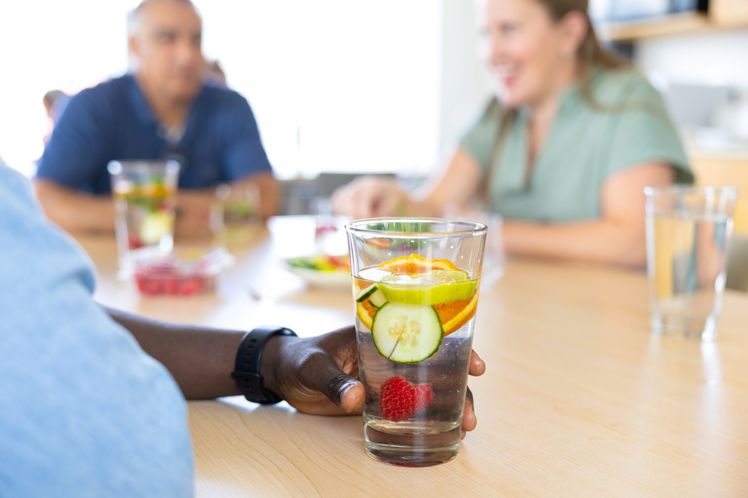 A person holds a glass of water with cucumber, orange, and raspberry; two others sit blurred in the background at a table.